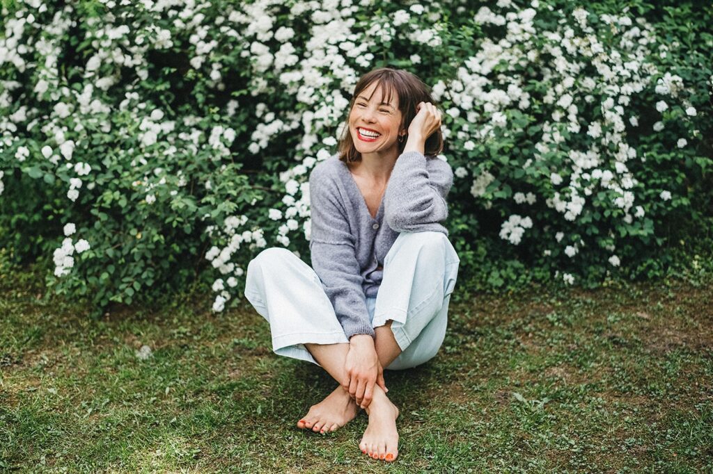 Eine Frau mit braunem Haar sitzt barfuß im Gras vor weiß blühenden Sträuchern, lächelt und berührt ihr Haar. Sie trägt einen hellvioletten Pullover und eine weiße Hose - aufgenommen von einem Familienfotografen aus dem Fotostudio Köln.