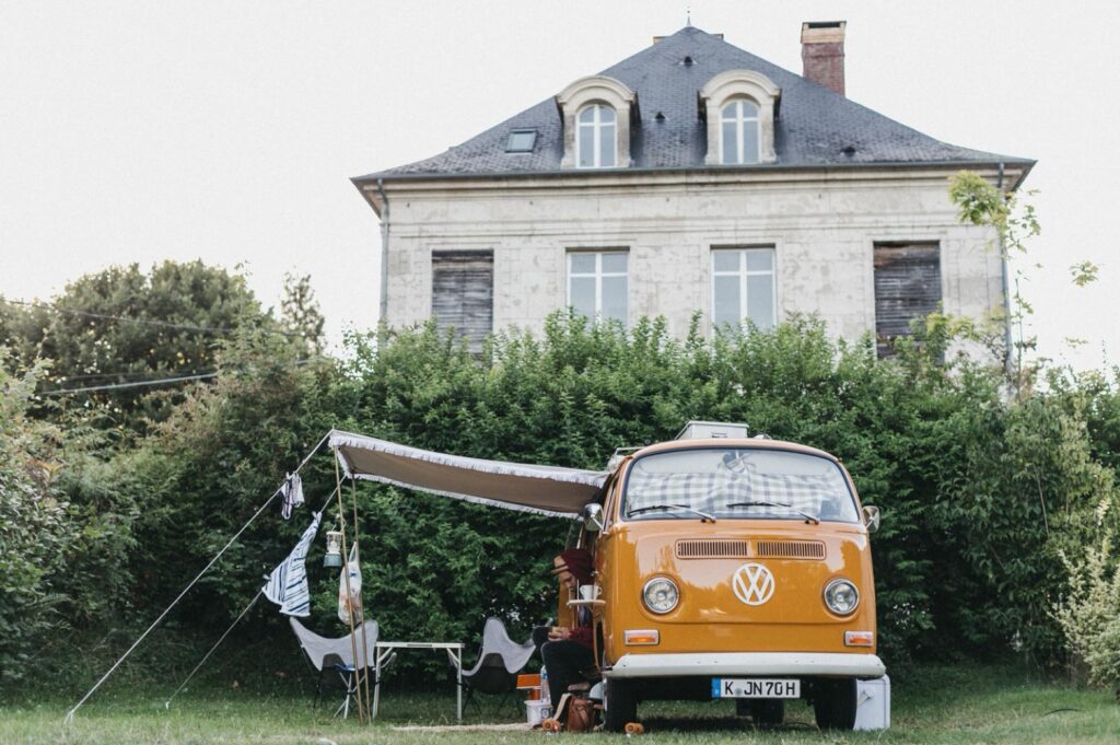Ein orangefarbener Oldtimer-Volkswagen mit Vorzelt parkt vor einem großen alten Steinhaus, das von einem Familienfotografen perfekt eingefangen wurde. Campingstühle und Kleidung sind in der Nähe aufgestellt, umgeben von viel Grün an einem sonnigen Tag.