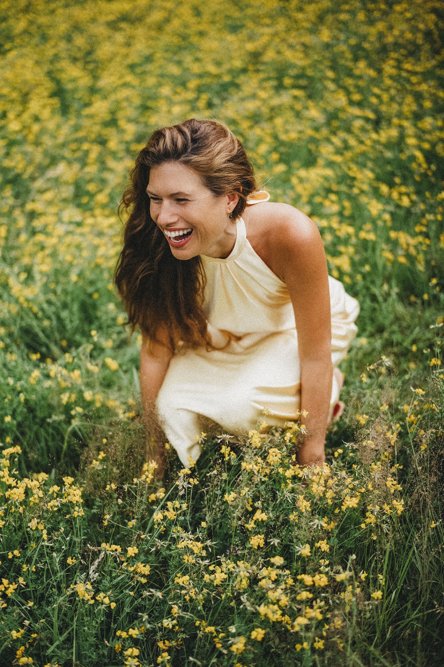 Eine junge Frau in einem cremefarbenen Kleid lacht, während sie in einem Feld mit gelben Wildblumen kniet, aufgenommen von einer talentierten Fotografin und Werbefotografin, umgeben von üppigem grünen Gras an einem sonnigen Tag.
