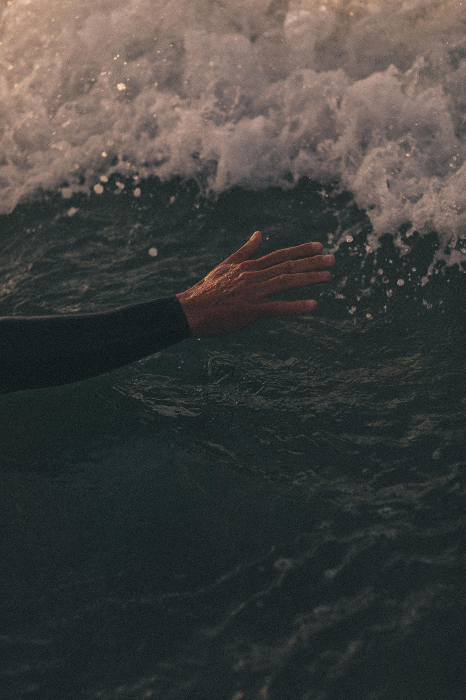 Die Hand einer Person, die teilweise von einem Neoprenanzug bedeckt ist, gleitet sanft über die Oberfläche des dunklen Meereswassers - eingefangen von einer talentierten Fotografin für das Fotostudio Köln, während im Hintergrund schäumende Wellen rollen.