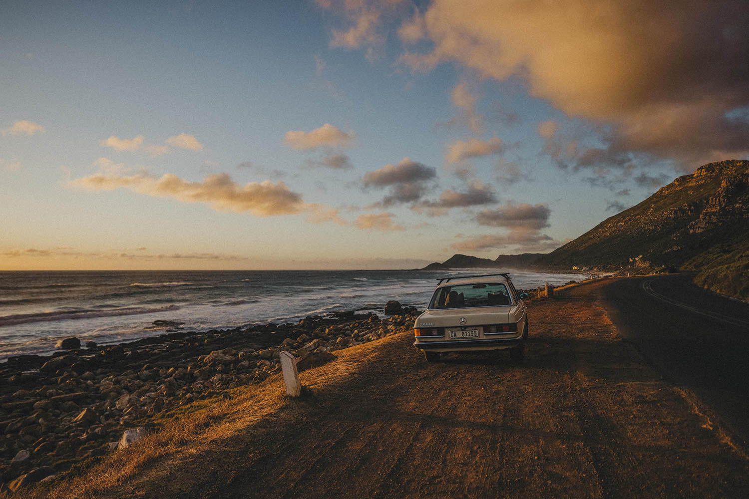 Ein Oldtimer parkt bei Sonnenuntergang an einer Küstenstraße mit Blick auf das Meer und die Wellen, die gegen die Felsen schlagen. Eingefangen von einer talentierten Fotografin, könnte diese Szene das Portfolio eines jeden Fotostudios Köln schmücken.