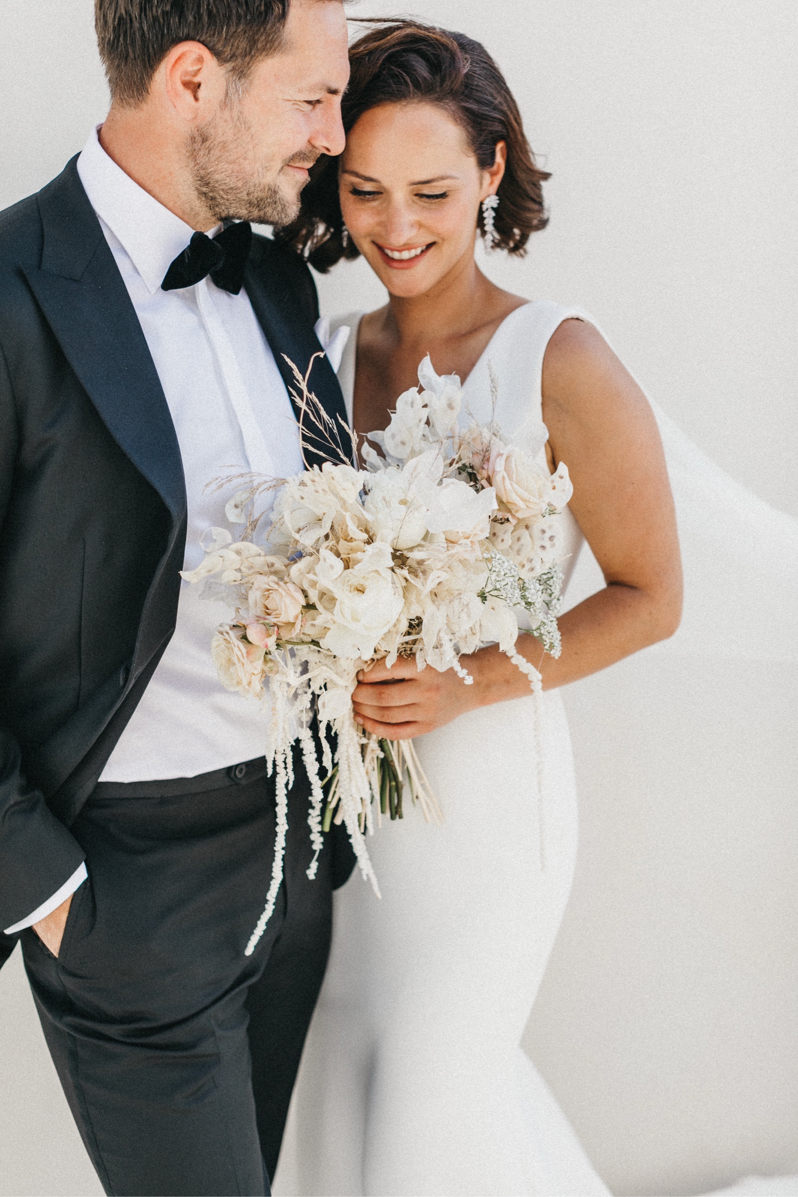 Eine Braut in einem weißen Kleid hält einen Strauß heller Blumen und lächelt mit geschlossenen Augen neben einem Bräutigam in einem schwarzen Smoking. Sie stehen dicht beieinander vor einem hellen Hintergrund, der die Romantik ihrer Hochzeit auf Sizilien einfängt.