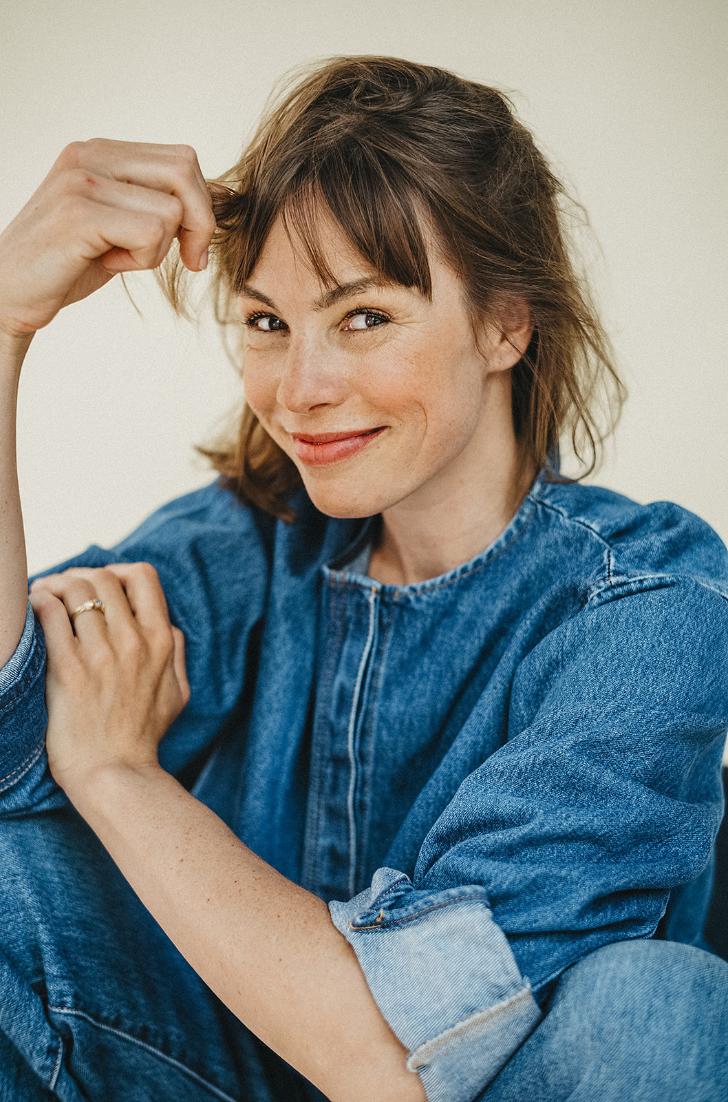 Eine lächelnde Frau mit hellbraunem Haar und Pony, die ein Jeanshemd mit hochgekrempelten Ärmeln trägt, sitzt mit einem Arm auf dem Knie vor einem schlichten Hintergrund in einem Fotostudio Köln.