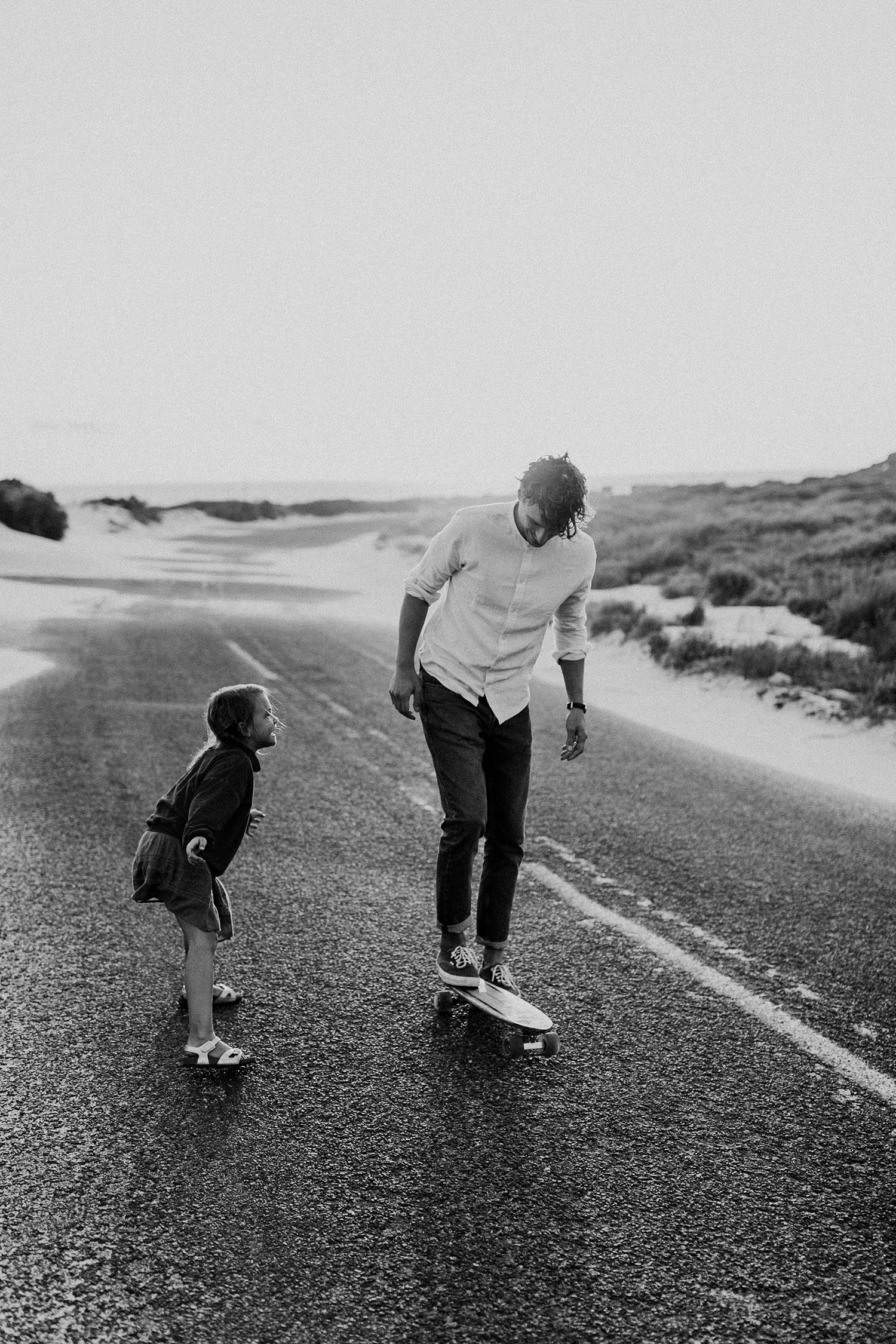 Ein Mann fährt mit dem Skateboard auf einer leeren Straße, während ein Kind neben ihm läuft, beide lächeln und genießen den Moment. Die Schwarz-Weiß-Aufnahme eines Familienfotografen zeigt Dünen und Grünflächen in einer malerischen Kulisse.