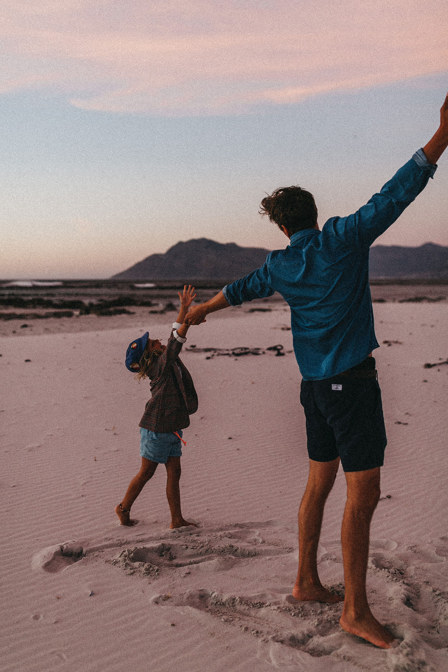 Ein Mann und ein Kind halten sich an den Händen und spazieren spielerisch über einen Sandstrand bei Sonnenuntergang, mit Bergen in der Ferne und einem zartrosa Himmel über ihnen - eingefangen von einem Familienfotografen mit einem Auge für herzliche Momente.