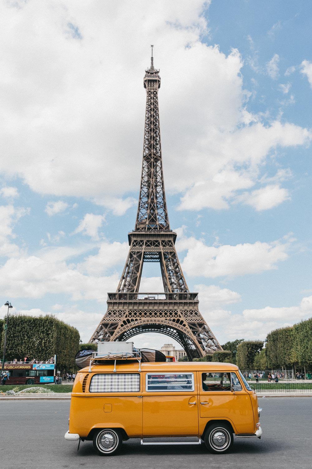 Ein leuchtend gelber Oldtimer-Van parkt an einem sonnigen Tag vor dem Eiffelturm in Paris, mit grünen Bäumen und einem blauen Himmel mit weißen Wolken im Hintergrund - die perfekte Inspiration für jeden Familienfotografen, der auf der Suche nach lebendigen Reisemomenten ist.